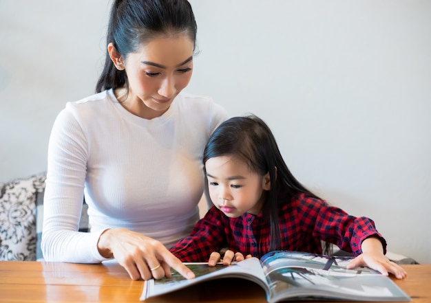 Uma mãe asiática está ensinando a filha a ler um livro durante o semestre na mesa da sala e tomando leite frio na mesa em casa. conceitos e atividades educacionais da família