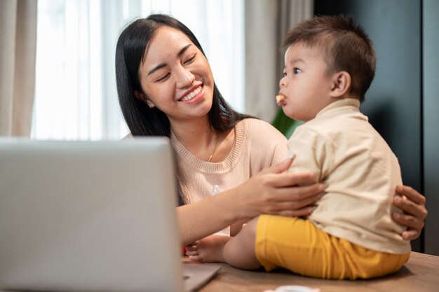 Uma mãe asiática feliz está brincando e alimentando seu filhozinho com lanches enquanto trabalha em seu escritório em casa