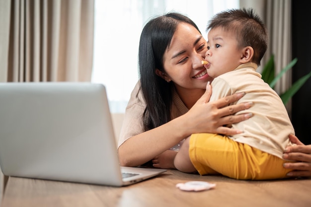 Uma mãe asiática feliz está brincando e alimentando seu filhozinho com lanches enquanto trabalha em seu escritório em casa