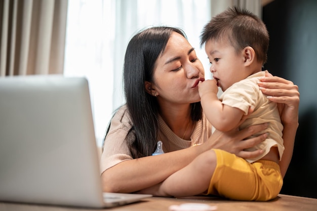 Uma mãe asiática feliz está brincando e beijando seu filhozinho bonito enquanto trabalha em seu escritório em casa