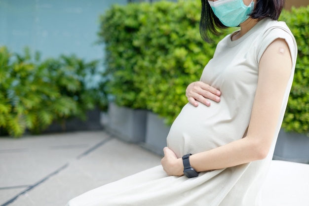 Uma mãe asiática grávida com um vestido longo está usando uma máscara e se sentando depois de ser vacinada.