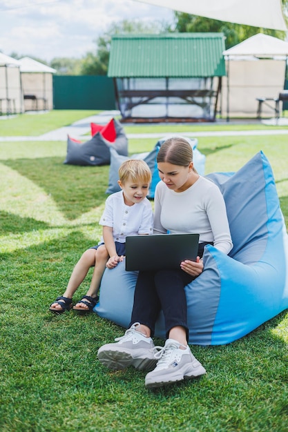 Uma mãe com um laptop seu filho sentado ao lado dela ao ar livre em um gramado verde Descanse e trabalhe com uma criança