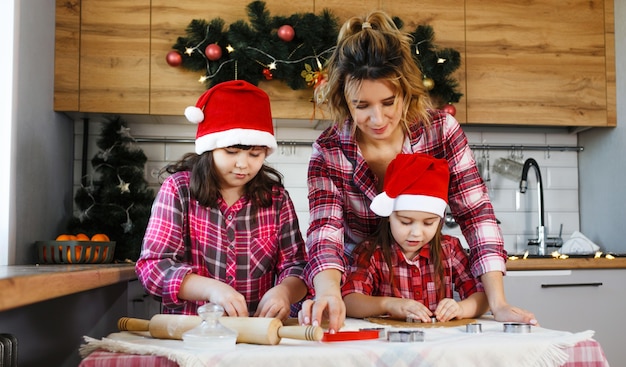 Uma mãe e duas filhas com camisas xadrez vermelhas e chapéus vermelhos aprendem a fazer biscoitos na cozinha decorada para o natal