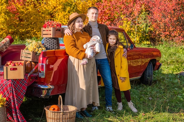 Uma mãe encorpada com um recém-nascido e filha e pai fica perto de um carro retrô vermelho carregado com caixas de legumes no contexto da natureza rural do outono