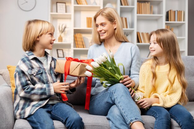 Foto uma mãe entusiasmada recebe um buquê e uma caixa de presentes de seu filho e filha todos aconchegados no sofá