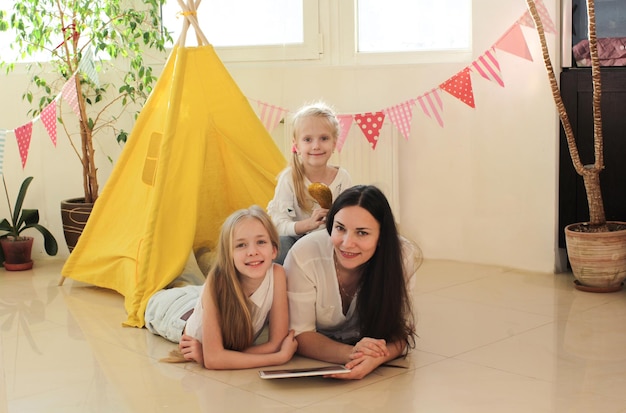 Uma mãe feliz com duas filhas se diverte deitada na sala de estar em uma tenda amarela.