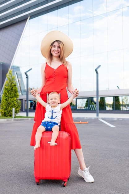 Uma mãe feliz com um filho bebê no aeroporto com uma mala vermelha sai em uma viagem ou férias no verão