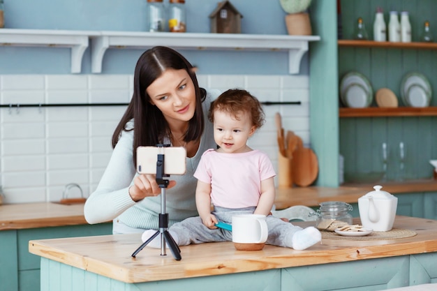 Uma mãe feliz com uma criança senta-se na cozinha à mesa e grava vídeo para o blog em um smartphone conectado a um tripé