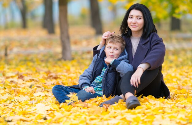 Uma mãe feliz e um filho sentado entre as folhas amarelas do outono no parque.