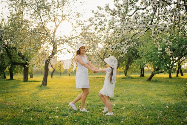 Uma mãe fofa de chapéu e vestido branco está andando pelo jardim da primavera com sua filha fofa