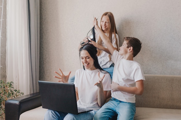 Uma mãe irritada se senta no sofá e trabalha em casa em um laptop, crianças ativas brincam com o cabelo da mãe, arruma o cabelo dela