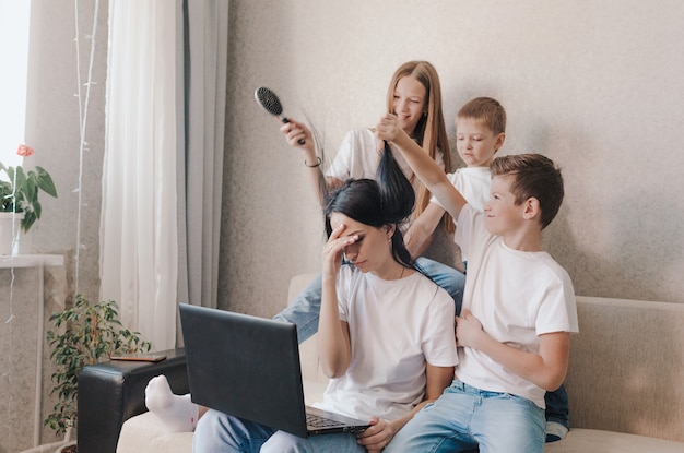 Uma mãe irritada se senta no sofá e trabalha em casa em um laptop, crianças ativas brincam com o cabelo da mãe, arruma o cabelo dela