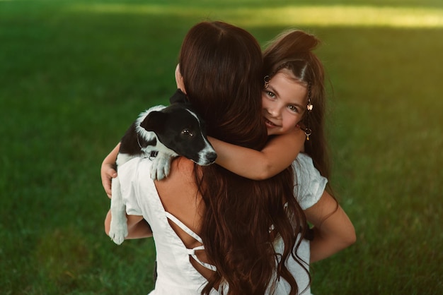 Uma mãe leva sua filha e um cachorrinho contra a grama verde no parque vista de trás família feliz amor por animais de estimação emoções de felicidade