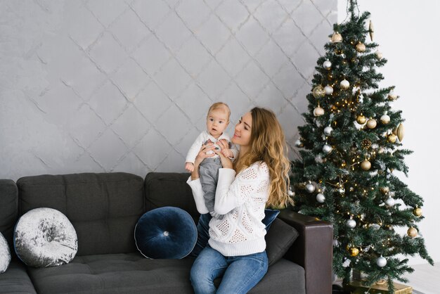 Uma mãe segura seu filho nos braços e sorri para ele enquanto está sentado perto de uma árvore de natal na sala de estar em sua casa