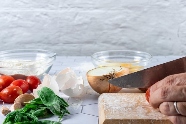 Uma mão cortando tomate entre ingredientes e utensílios de cozinha