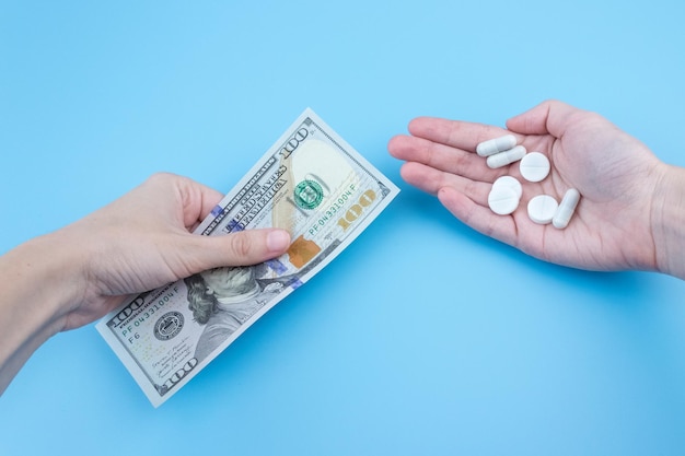 Uma mão dá cem dólares para uma mão em uma luva médica com um comprimido em um fundo azul. Aumento do preço dos medicamentos. Negócio farmacêutico