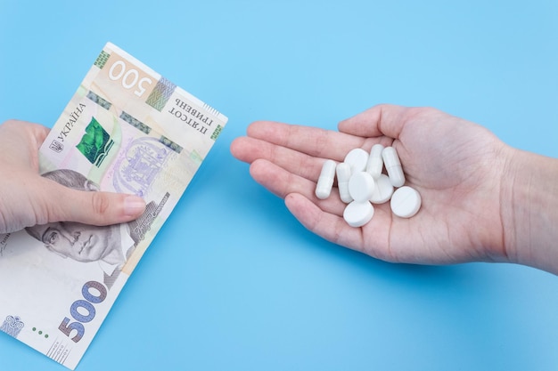 Uma mão dá uma conta de 500 hryvnia a uma mão em uma luva médica com um comprimido em um fundo azul. aumento do preço dos medicamentos