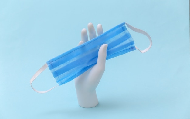 Uma mão de manequim branco segura uma máscara médica sobre um fundo azul. Conceito de minimalismo de medicina