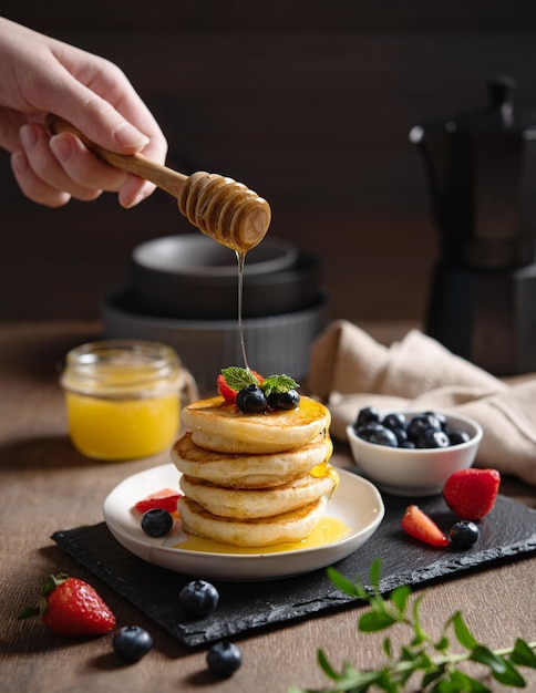 Uma mão derrama mel em panquecas americanas clássicas em uma pilha com frutas frescas em um fundo marrom embaçado O conceito de um café da manhã saudável, delicioso e nutritivo