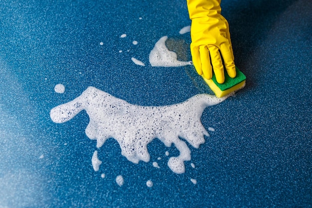 Uma mão em uma luva de borracha amarela lava uma parede com uma esponja molhada e espumosa azul espuma de fundo azul espaço de cópia conceito de limpeza tarefas domésticas limpeza de primavera