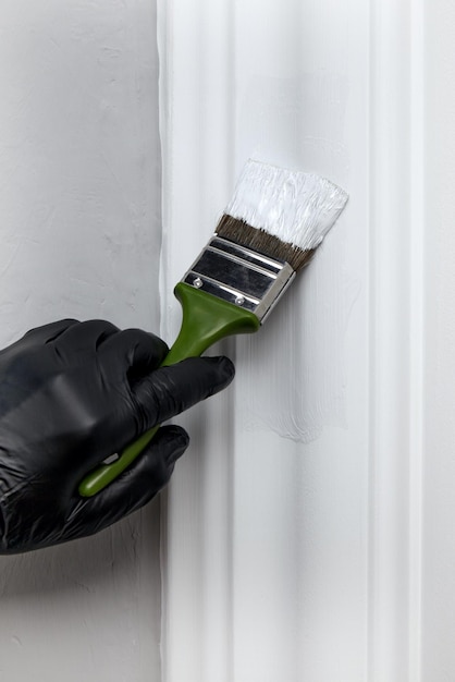 Uma mão em uma luva de borracha preta pinta uma moldura de porta decorativa com tinta branca com pincel