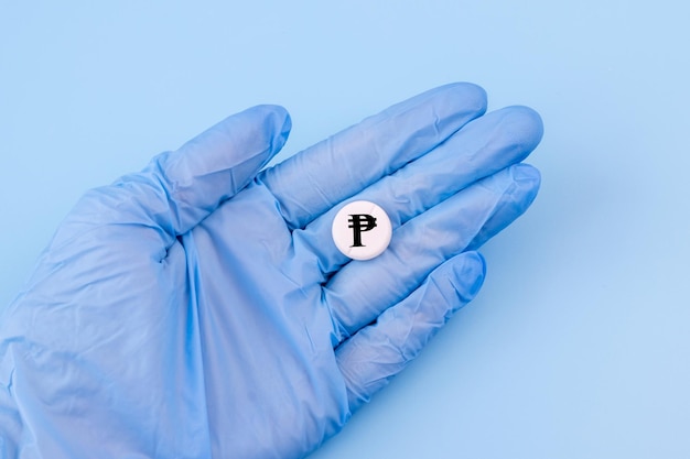 Uma mão em uma luva médica segura uma pílula com um sinal de pesos em um fundo azul Remédio caro nas Filipinas