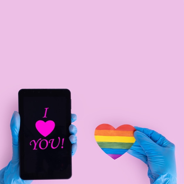Foto uma mão em uma luva protetora segura um smartphone com a inscrição eu te amo, e uma mão em uma luva médica azul segura uma forma de coração de papel do arco-íris em um fundo rosa. conceito lgbt