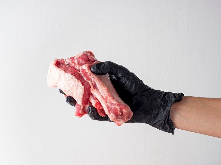 Uma mão enluvada negra segura um pedaço de carne crua contra um fundo claro.