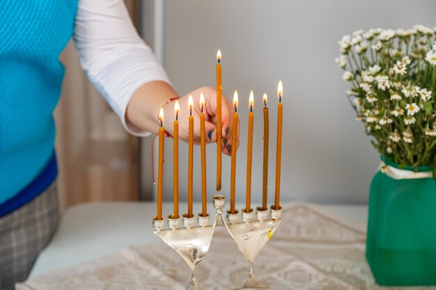 Uma mão feminina acende velas em Hanukkah na mesa ao lado de um vaso de flores. Foto horizontal