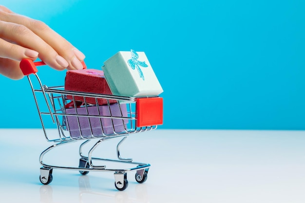 Uma mão feminina empurrando o carrinho de compras com caixas de presente em estúdio em fundo azul