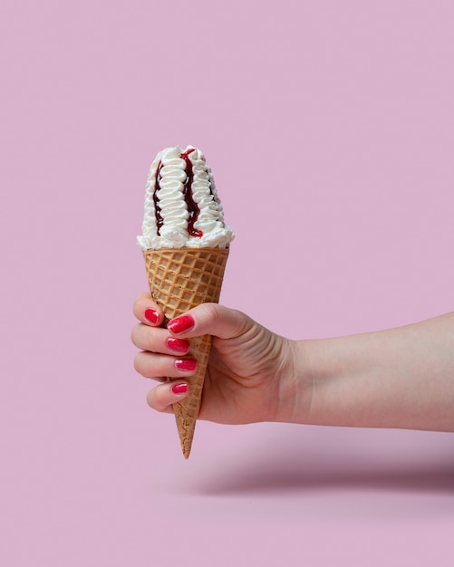 Uma mão feminina segura uma casquinha de sorvete com geleia de cereja em um copo de waffle em um fundo rosa.