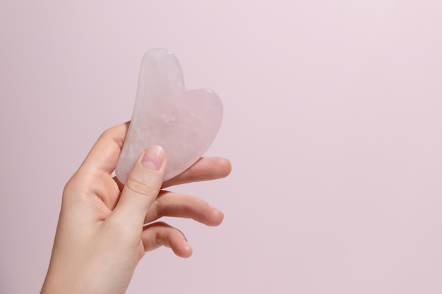 Uma mão feminina segurando o massageador facial gua sha no fundo rosa