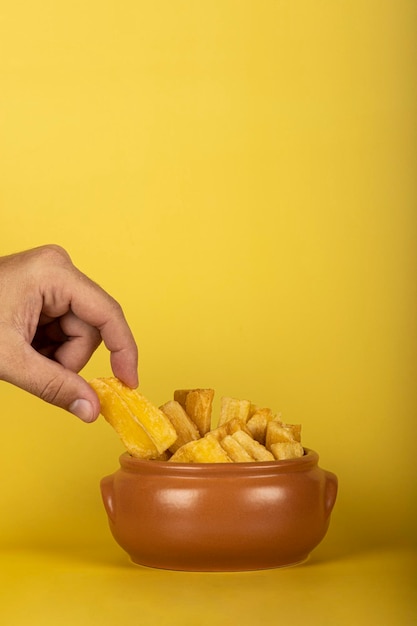 Foto uma mão pegando uma mandioca frita em cima de uma porção de mandiocas fritas sobre um fundo amarelo