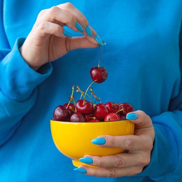 Foto uma mão segura uma tigela amarela com cerejas a outra segura uma baga pelo caule