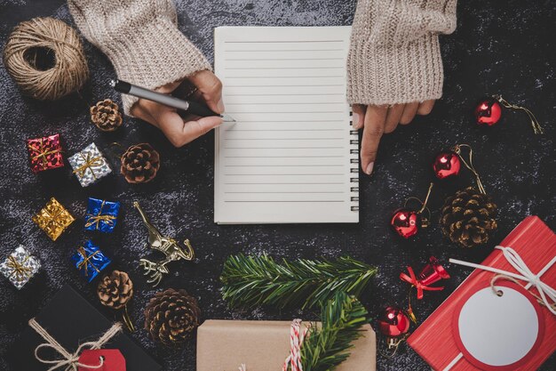 Uma mão segurando uma caneta para escrever em um caderno com óculos e pequenos presentes em cimento preto