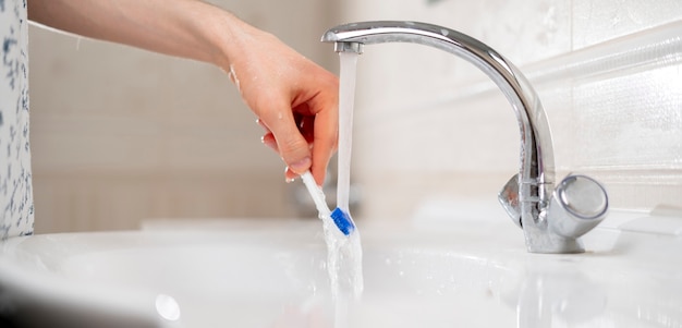 Uma mão segurando uma escova de dentes branca e lavando-a debaixo d'água na pia do banheiro