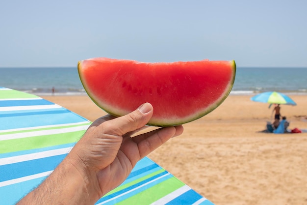 Uma mão segurando uma fatia de melancia, na praia. Conceito da maneira natural de se refrescar..