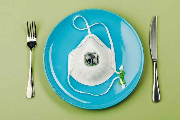 Uma máscara médica em um prato vazio com garfo e faca em um fundo verde