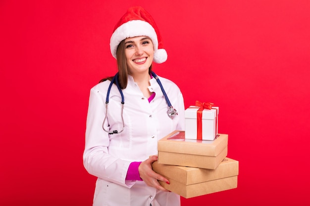 Uma médica com um chapéu de Papai Noel segura caixas com presentes em um fundo vermelho Parabéns do médico aos pacientes