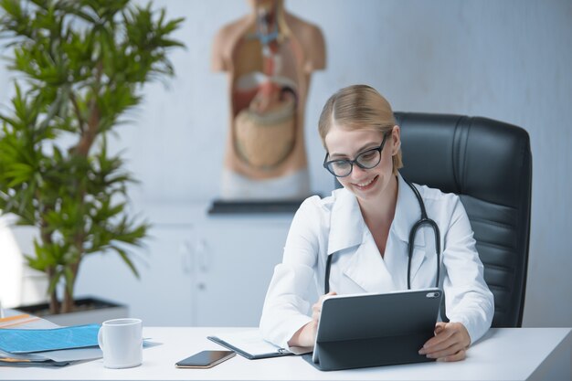 Uma médica com um estetoscópio no pescoço trabalha em uma mesa com um tablet em um escritório bem iluminado