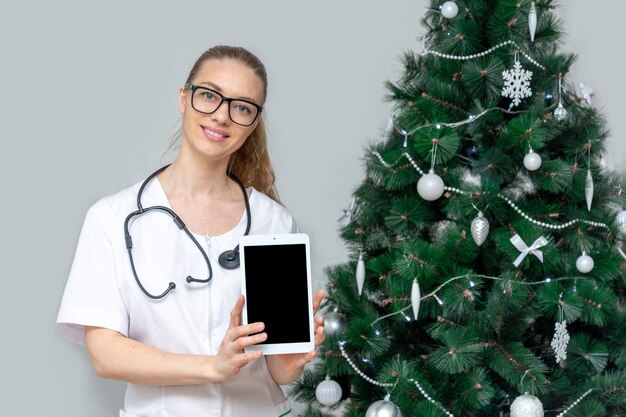 Uma médica com uma máscara médica protetora segura um tablet no fundo de uma árvore de Natal.