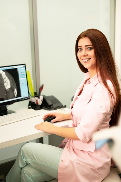 Uma médica dentista está sentada à mesa, em um computador, uma tomografia computadorizada da mandíbula. O médico está vestido com roupas profissionais.