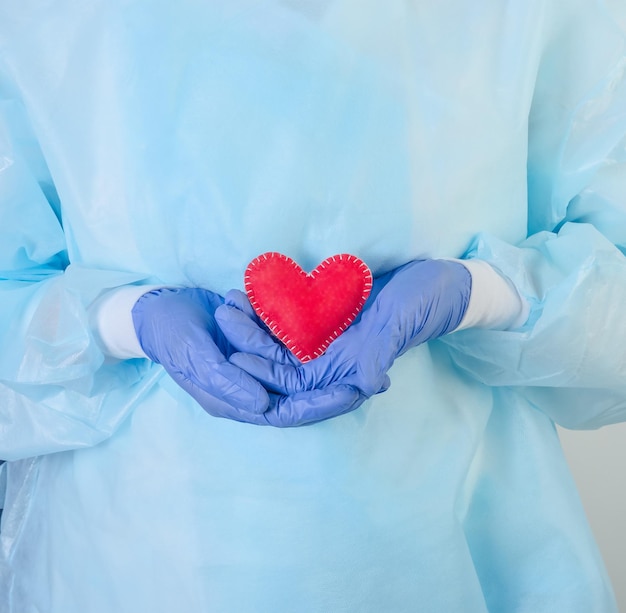 Uma médica em um traje de proteção segura um coração vermelho nas mãos em luvas em um fundo branco