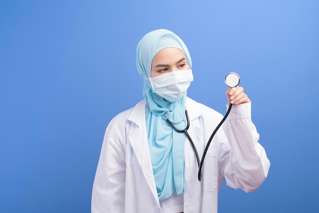 Uma médica muçulmana com hijab usando uma máscara cirúrgica sobre estúdio de fundo azul.