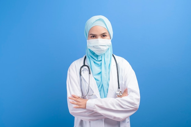 Uma médica muçulmana com hijab usando uma máscara cirúrgica sobre uma parede azul.