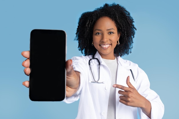 Uma médica negra milenar positiva com jaleco branco aponta o dedo para um smartphone com tela em branco