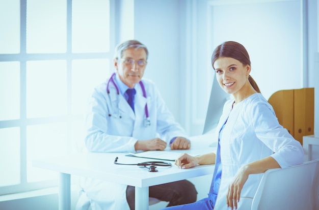 Uma médica sorrindo para a câmera com seu colega no fundo do consultório de um hospital