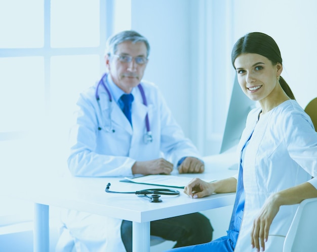 Uma médica sorrindo para a câmera com seu colega no fundo do consultório de um hospital