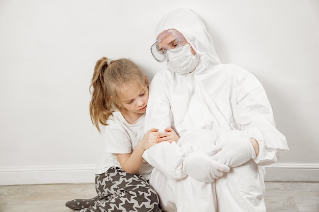 Uma menina abraça um médico em uma roupa protetora branca, máscara, óculos e luvas. zona vermelha. Paciente bebê. Gratidão. medicina durante uma pandemia. mamãe é médica