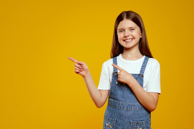 Uma menina adorável vestindo um vestido de jeans sorri enquanto aponta com confiança com os dedos indicadores
